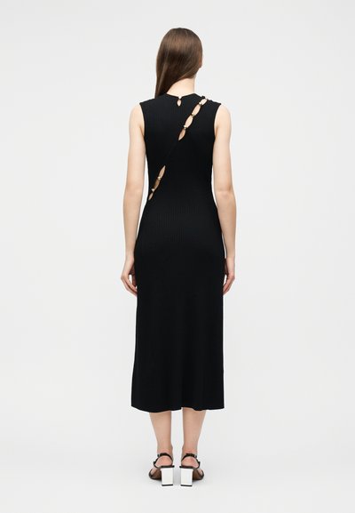 Femme portant une robe midi noire sans manches avec une découpe diagonale et des anneaux dorés dans le dos, debout, chaussée de talons blocs noirs et blancs.