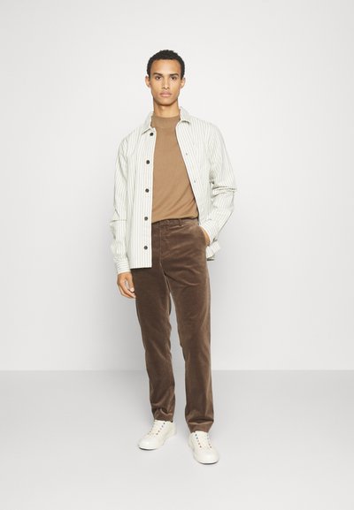 Striped light green button-up shirt, brown t-shirt, brown corduroy pants, and white sneakers. Model standing against a plain background.