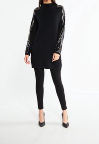 Robe en maille noire à manches longues ornée de détails en sequins et d'accents métalliques, portée avec des leggings noirs et associée à des escarpins noirs à bouts pointus.