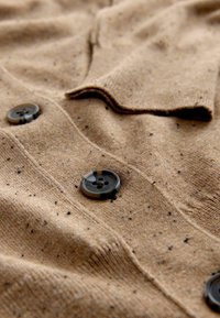 Maglione lavorato a maglia color marrone chiaro con finiture texture, caratterizzato da piccole macchie scure, un colletto e due bottoni neri.