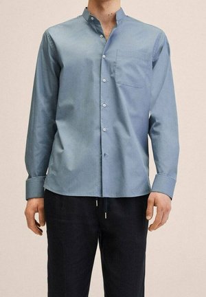 Uomo che indossa una camicia azzurro chiaro a maniche lunghe con colletto alla cinese e pantaloni neri, in piedi davanti a uno sfondo neutro.
