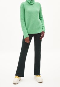 Grön stickad turtleneck-tröja med ribbad textur, kombinerad med mörkgröna utsvängda byxor och vita sneakers med gula skosnören.