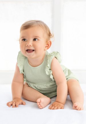 Grenouillère bébé vert clair avec manches à volants, présentant une texture lisse et un motif de petits pois. Le bébé a les cheveux courts et bouclés et sourit.