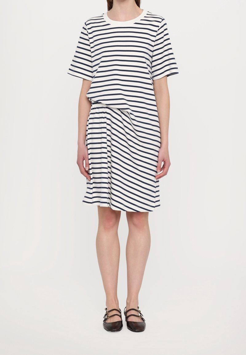 Femme portant une robe blanche à manches courtes avec des rayures horizontales noires, une taille élastique et des chaussures Mary Jane noires.