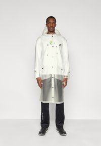 Λευκό αδιάβροχο παλτό με διαφανές κάτω τμήμα, μαύρα κουμπιά και κουκούλα, με πράσινη σχεδιαστική λεπτομέρεια στο στήθος.