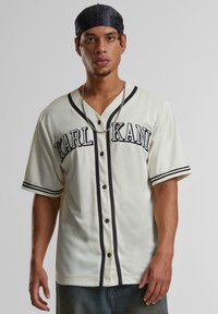 Tricou de baseball crem cu accente negre, buton în față, mâneci scurte și inscripția "KARL KANI" imprimată pe piept. Material moale, texturat.