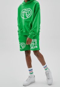 Hombre vestido con una sudadera verde y pantalones cortos a juego con impresiones gráficas y de paz en blanco, zapatillas blancas y calcetines blancos con rayas azules y verdes.