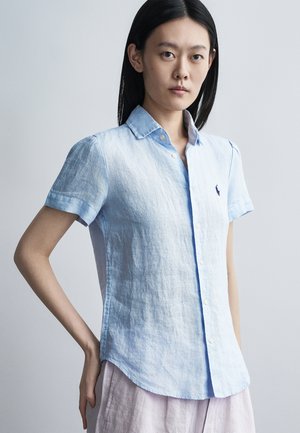 Jeune femme aux cheveux noirs et raides porte une chemise bleu clair à manches courtes boutonnée et un pantalon clair, debout avec une main sur la hanche.