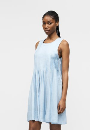 Kobieta z warkoczami, ubrana w bezrękawnikową, jasnoniebieską plisowaną sukienkę, stojąca na tle jednolitego jasnego tła.