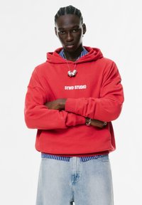 Jonge man met gevlochten haar, gekleed in een rode hoodie met de tekst "STWD STUDIO", een blauw gestreept shirt eronder, lichte jeans en een zilveren halsketting.