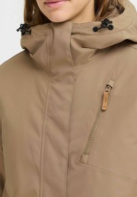 Braune wasserdichte Jacke mit hohem Kragen, verstellbaren Kordeln und einer seitlichen Reißverschlusstasche mit einem Holz-Zuggriff. Glatte Stoffstruktur.