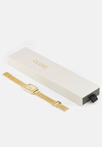 Montre de couleur or avec un cadran rectangulaire et un bracelet en maillons métalliques, accompagnée d'une boîte crème avec un logo doré.
