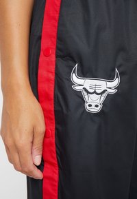 Czarne sportowe spodnie z czerwonymi bocznymi paskami i zatrzaskami, z białym wyhaftowanym logo byka na udzie. Gładki, lekki materiał.