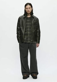 Young person with long hair wearing a black leather jacket, green plaid shirt, wide dark pants, and black shoes, standing against a plain background.