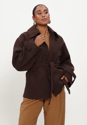 Woman wearing brown belted coat and tan pants, standing with one hand near collar and the other in pocket against plain background.