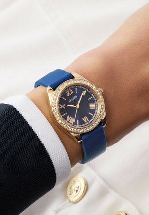 Blauwe horlogeband van siliconen met een kast in goudkleur, een marineblauwe wijzerplaat, Romeinse cijfers en kristalaccenten rondom de lunette. Contrast met een gestreepte mouw.