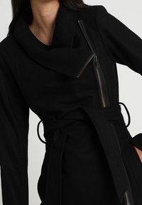 Černý kabát zavinovacího střihu s širokým límcem, diagonálním zipem a páskem v pase. Vyroben z měkké, texturované látky.