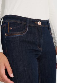 Jean en denim bleu foncé avec des coutures contrastantes en orange et bleu, doté d'une taille mi-haute, de poches avant et d'une fermeture à bouton en cuivre.