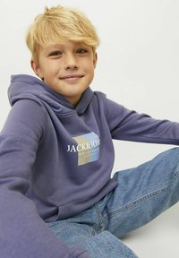 Paarse hoodie met een voorkant grafisch ontwerp met blauwe en beige vormen, gecombineerd met blauwe denim jeans, zittend tegen een neutrale achtergrond.