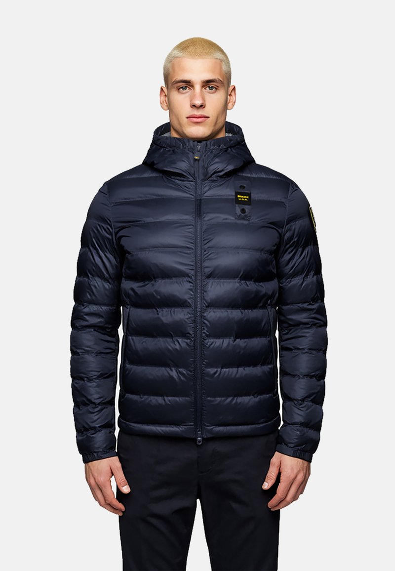 Navy blue puffer jacket with a zippered front, hood, and visible horizontal stitching. Features a logo patch on the chest and button details on the side.