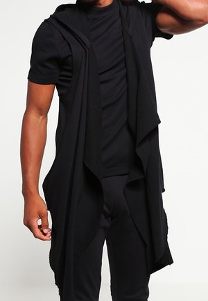 Man draagt een gelaagde zwarte outfit, bestaande uit een T-shirt met korte mouwen en een lang, mouwloos openvest met een gedrapeerde kraag.