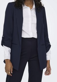 Blazer bleu marine en tissu léger avec une coupe ajustée, des revers crantés et des manches retroussées, accompagné d'une chemise blanche à boutons.