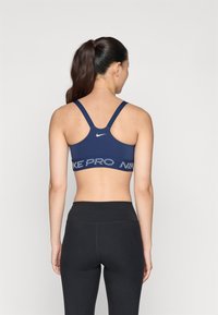 Jūras spēku sporta krūšturis ar platu melnu joslu, kurā redzams teksts "NIKE". Regulatora tipa aizmugure, gluda auduma struktūra, minimālistisks logs aizmugurē.