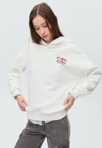 Felpa bianca con cappuccio e logo ricamato rosso, vestibilità ampia, polsini e orlo a costine. Il materiale sembra morbido e strutturato.