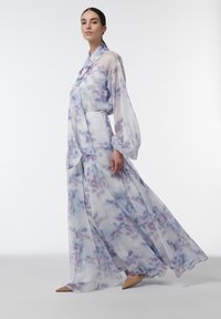 Mujer con un vestido largo, fluido y translúcido con un estampado de acuarela en tonos pastel morado y azul, tacones color beige y el pelo recogido, caminando hacia la izquierda.