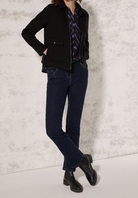 Veste en laine noire avec poches avant, portée sur une chemise violette à motifs. Associée à un jean bleu foncé et des bottines noires.