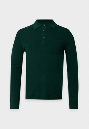 Jeune homme debout, mains dans les poches, portant un polo à manches longues vert foncé, un jean gris à jambes larges et des baskets marron sur fond blanc.