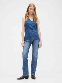 Young woman with long red hair wearing a sleeveless blue denim buttoned top, blue jeans, and black ankle boots standing against a plain background.