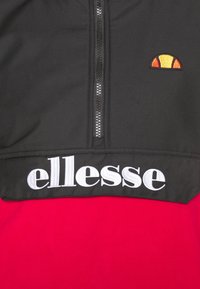 Sudadera con cremallera en negro y rojo, con un logo bordado en naranja y amarillo y el nombre de la marca "ellesse" en blanco en la banda inferior.