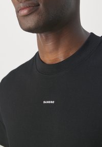 Gros plan d'une personne portant un T-shirt noir avec le mot "SANDRO" imprimé en petites lettres blanches sur la poitrine.