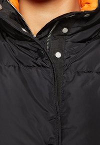 Doudoune noire avec une doublure intérieure orange, comportant une fermeture éclair et des boutons-pression argentés. Tissu texturé et conception isolée.