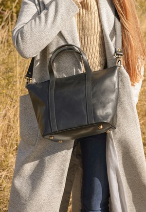 Person in beige sweater and gray coat carrying black leather handbag outdoors in sunlight.