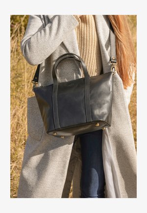 Osoba v béžovom svetri a šedej bunde nesúca čiernu koženú kabelku vonku na slnku.