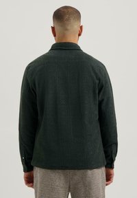 Man met kort haar in een donkergroene, gestructureerde blouse met lange mouwen en een geruit broek, staande met zijn rug naar de camera tegen een effen achtergrond.