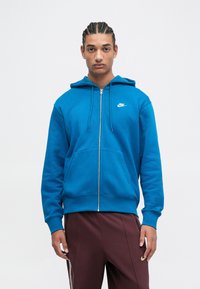 Sudadera azul con cremallera hecha de mezcla de algodón, que cuenta con un bolsillo canguro, capucha con cordón y un pequeño logo blanco de Nike en el pecho.