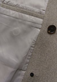 Beżowy wełniany płaszcz z widoczną gładką szarą podszewką, z przeszywanymi kieszeniami i dwoma ciemnymi guzikami na wierzchniej tkaninie.