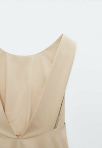 Beżowa sukienka bez rękawów z głębokim dekoltem na plecach, z szerokimi ramiączkami i gładkim, lekkim materiałem. Prosty fason bez widocznych wzorów.