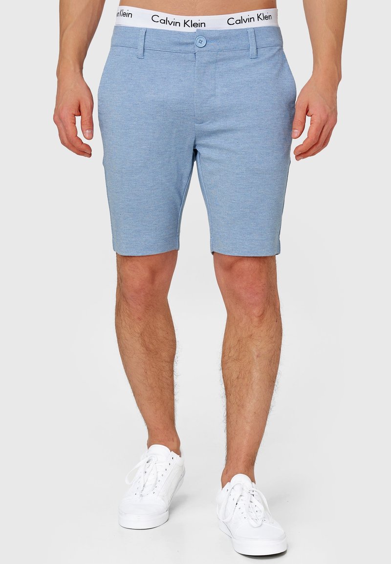 Short bleu clair cintré avec bouton et passants de ceinture, porté avec un sous-vêtement à ceinture blanche Calvin Klein et des baskets blanches.