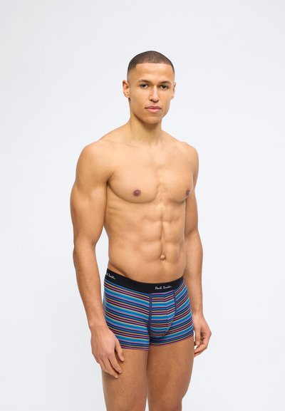 Μυώδης άνδρας με κοντά μαλλιά, που φοράει μποξεράκια με ρίγες σε μπλε, πορτοκαλί και πράσινο με μαύρη μέση, στέκεται μπροστά σε ένα απλό λευκό φόντο.