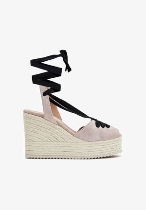 Sandalia cuña tipo esparto beige con suela trenzada y lazos negros que se atan alrededor del tobillo.