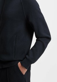 Sweat-shirt zippé bleu marine avec une texture lisse, des poignets côtelés et des poches latérales. Présente un design ajusté et des coutures plates pour un confort supplémentaire.