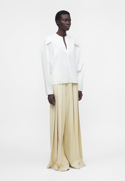 Femme portant un haut blanc à manches longues avec de larges épaules et un pantalon beige plissé à jambes larges, debout devant un fond blanc uni.