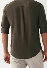Chemise vert foncé à manches longues avec manches retroussées et ourlet incurvé. Texture lisse, détail dos avec boutons, et fentes latérales.