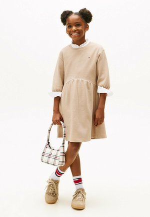 Beiges Sweatshirt-Kleid mit ausgestelltem Rock und weißem Rüschensaum. Braune Sneaker, eine gemusterte Tasche und gestreifte Socken runden das Outfit ab.