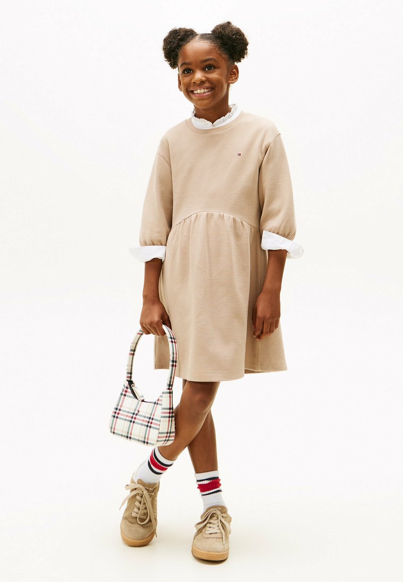 Robe-sweat beige avec une jupe évasée et un col blanc à volants. Des baskets marron, un sac à motifs et des chaussettes rayées complètent la tenue.