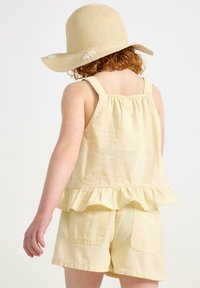 Vaaleankeltainen pellavapaita, jossa on röyhelöhelma ja säädettävät olkaimet, yhdistettynä samanvärisiin shortseihin, joissa on kaksi takata pockets ja yksinkertainen ompelu.
